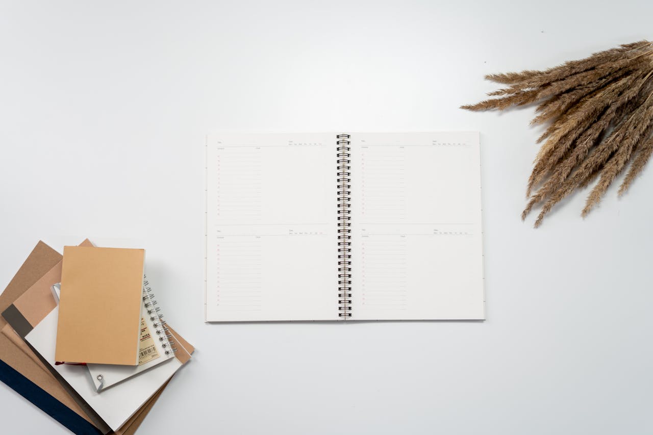 Open notebook with planner pages, surrounded by notebooks and pampas grass on a white background.
